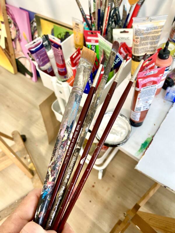 Mano sosteniendo pinceles usados delante de los tubos de pintura del estudio